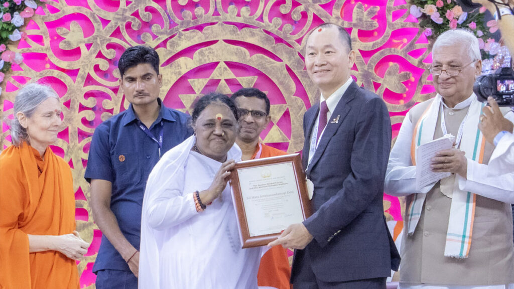 Amma holds her hand on the award next to a man in a suit who is awarding it to her