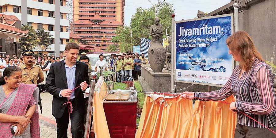 A plaque commemorating the Jivamritam collaboration is unvelied outside b a woman with others standing by. AIMS Hospital is in the background.