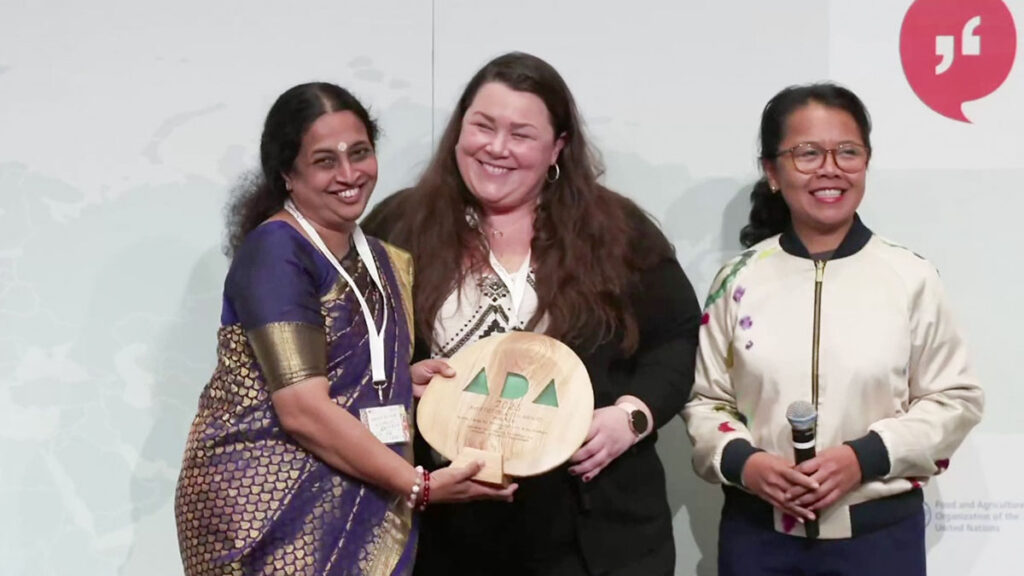 Dr Maneesha Vinodini Ramesh happily receiving the award on stage with two other people. Smiles all around.
