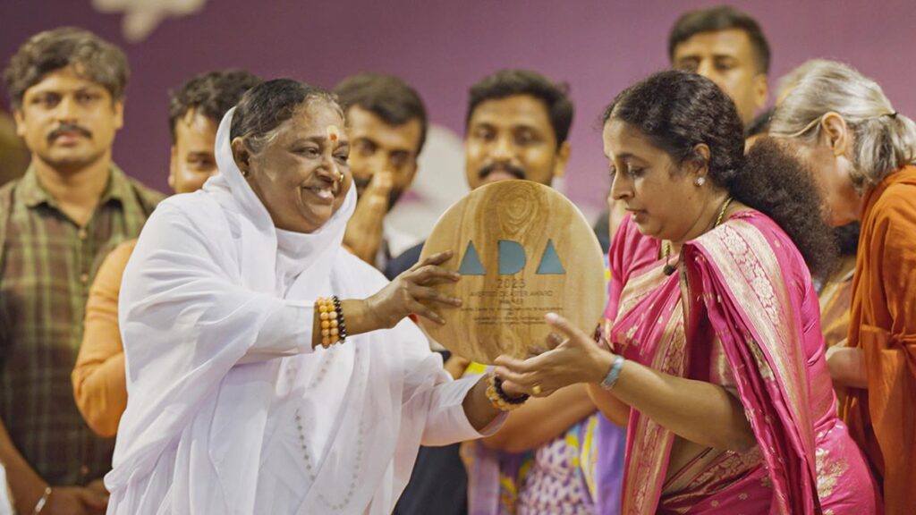 Amma, wearing a white sari, holding the award plaque, with Dr. Maneesha, who is dressed in a red and gold sari, and other team members looking on smiling