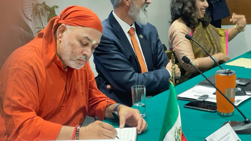 Swami Dayamritananda seated and writing at green cloth table with Mexico-Republic of India Friendship Group.