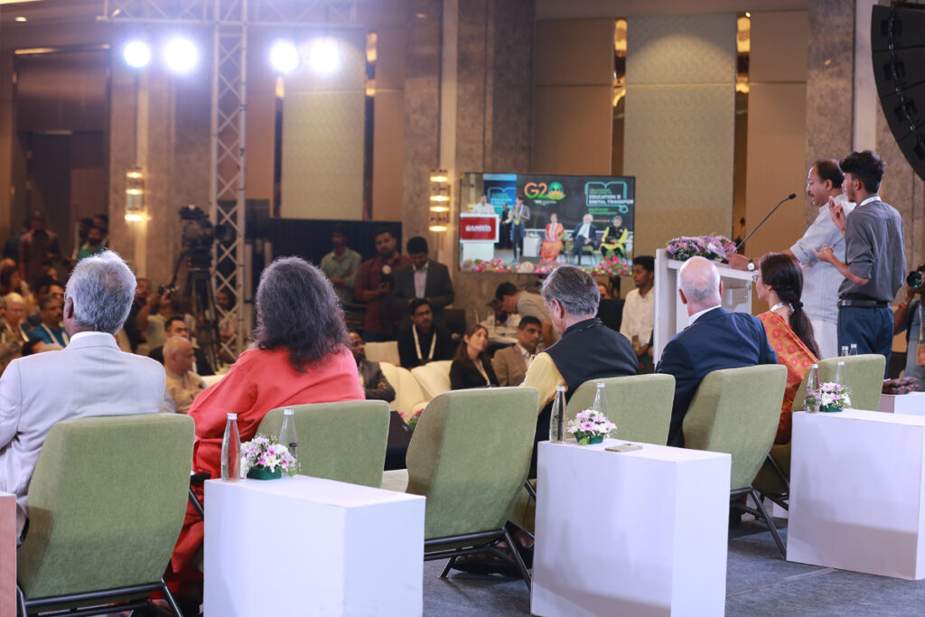 From behind the stage looking out, dignitaries sit in green cloth chairs on the stage as all listen to  Sri V Muraleedharan speak. 