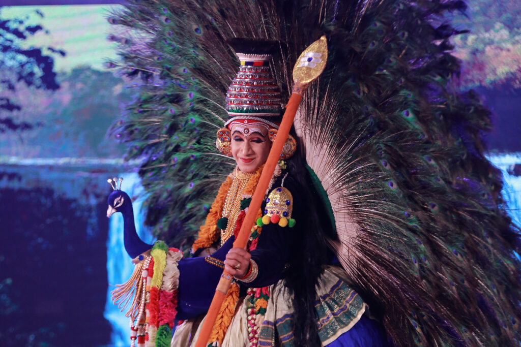 Classical dancer with a sublime smile as Sri Krishna, adornedfrom his back side with huge spray of peacock feathers, holding a golden bow with a bright blue costume and orange garland.