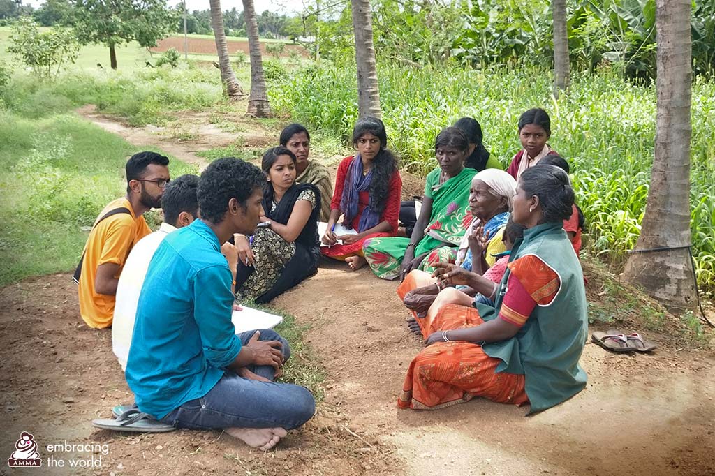 Students sit on the ground in a circle with villagers