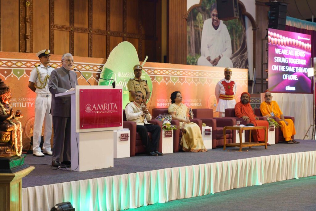 dignitaries seated on a low rise stage while Hon. Kerala Governor (Retd.) Justice P. Sadasivam stands at a podium.