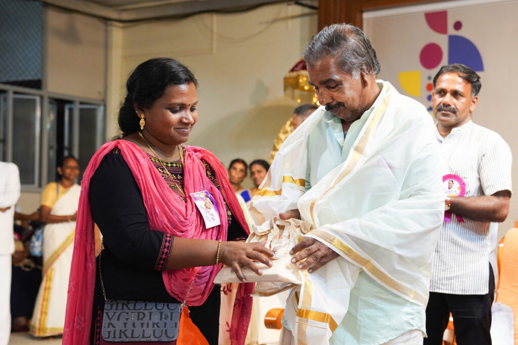 A woman smiles and adorns an older alumni man with a traditional Kerala cloth.