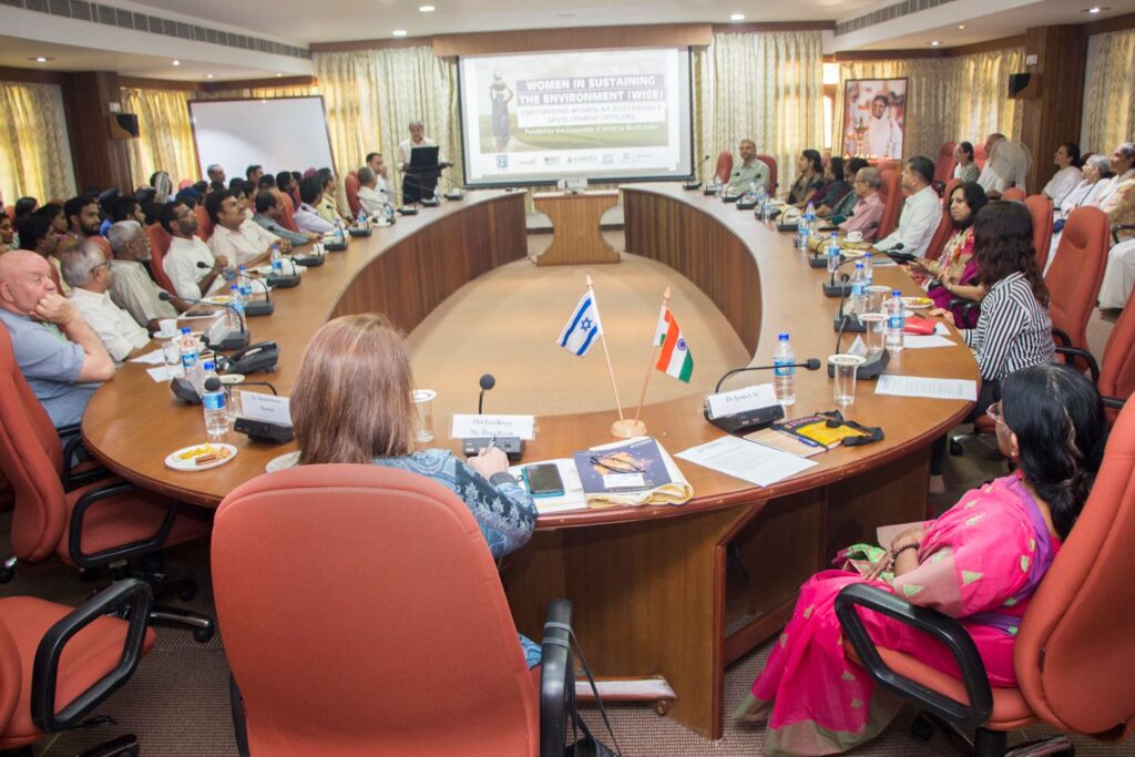 A large meeting takes place around an oval table in front of a screen