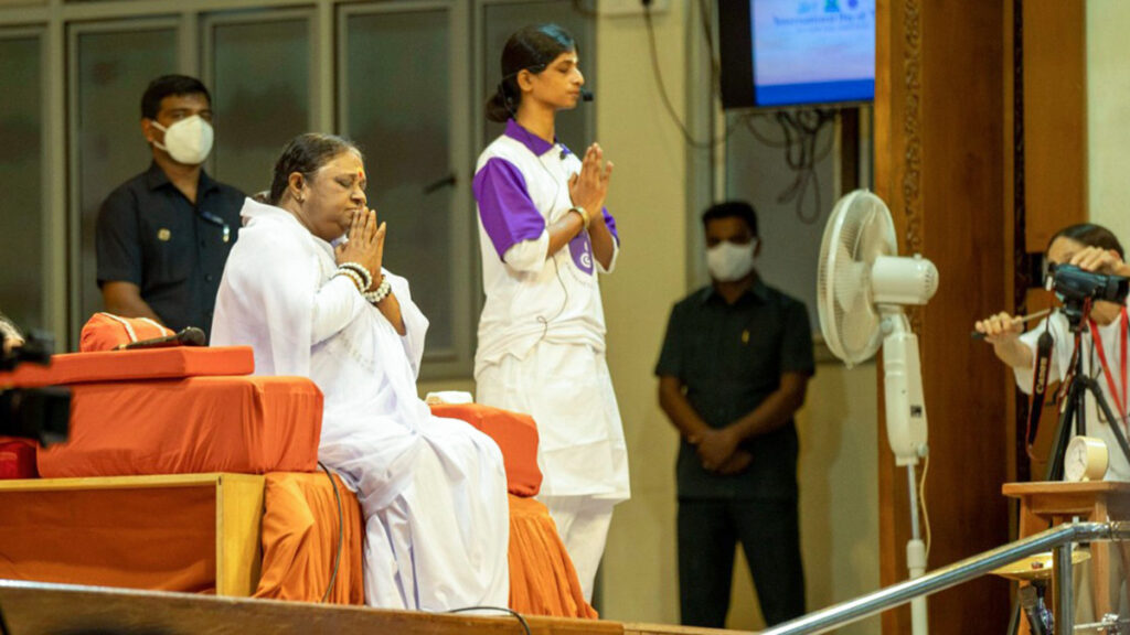 Amma sitting on stage next to female demonstrator holding standing yoga pose with palms together