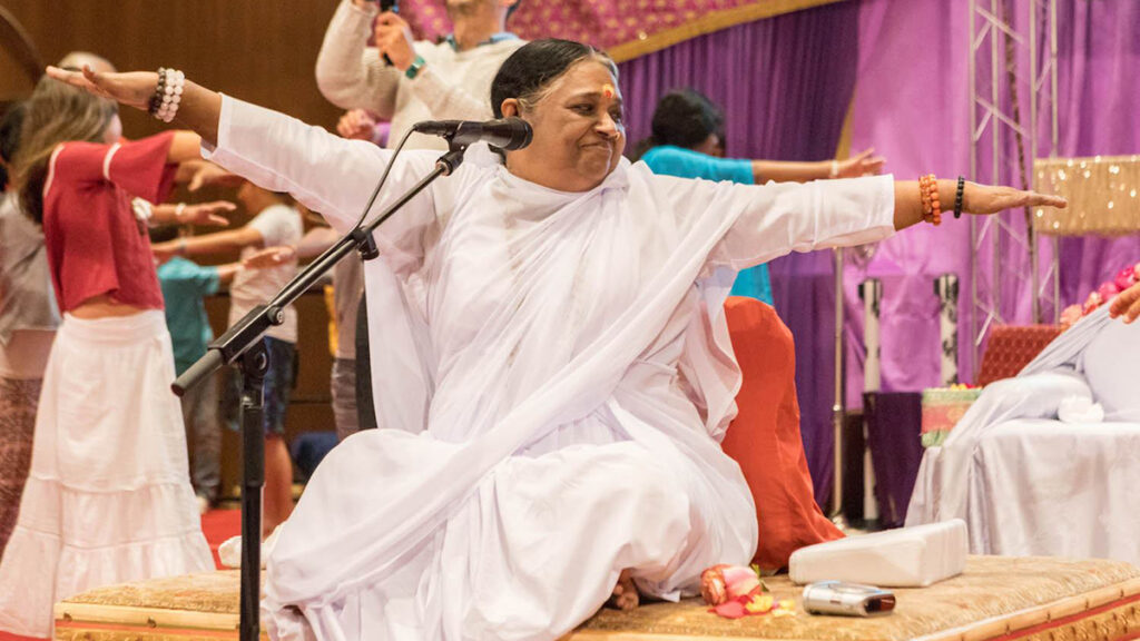 Amma seated on stage demonstrating a yoga pose with her arms outstretched to the sides